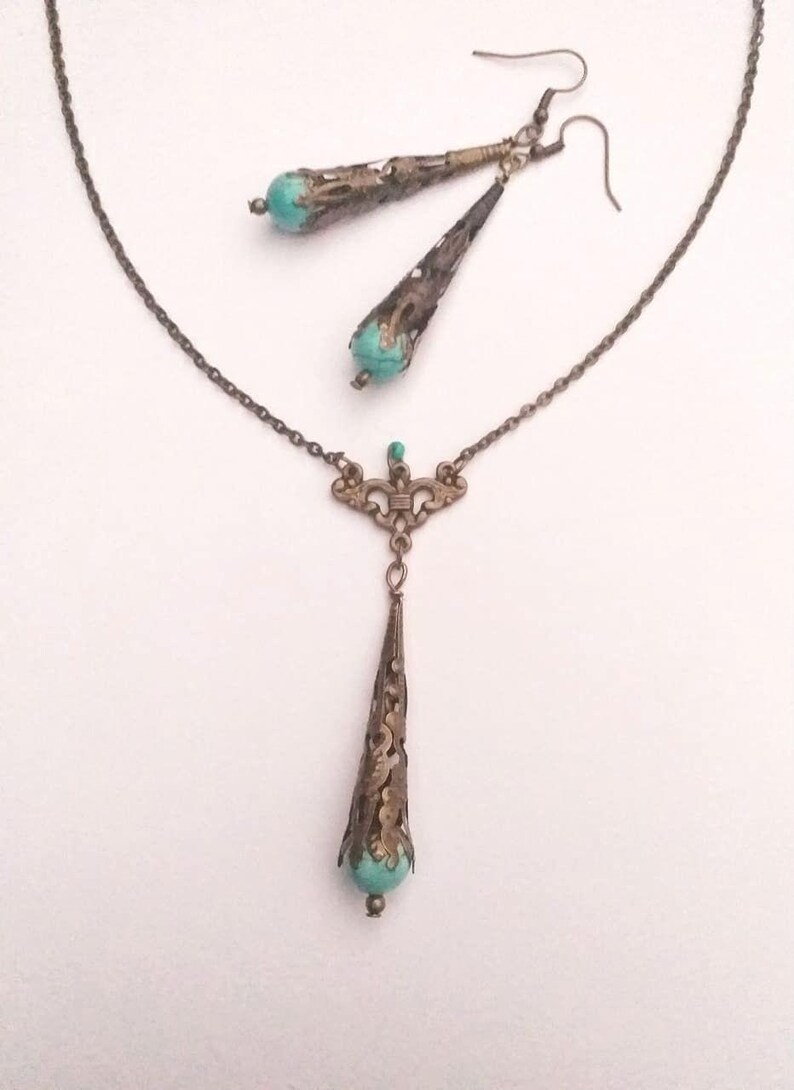 Puede incluir: Un conjunto de collar y pendientes de color turquesa y bronce con un delicado dise&ntilde;o de filigrana. El colgante tiene forma de l&aacute;grima con una piedra turquesa en la parte inferior. Los pendientes tambi&eacute;n tienen forma de l&aacute;grima con piedras turquesas en la parte inferior.