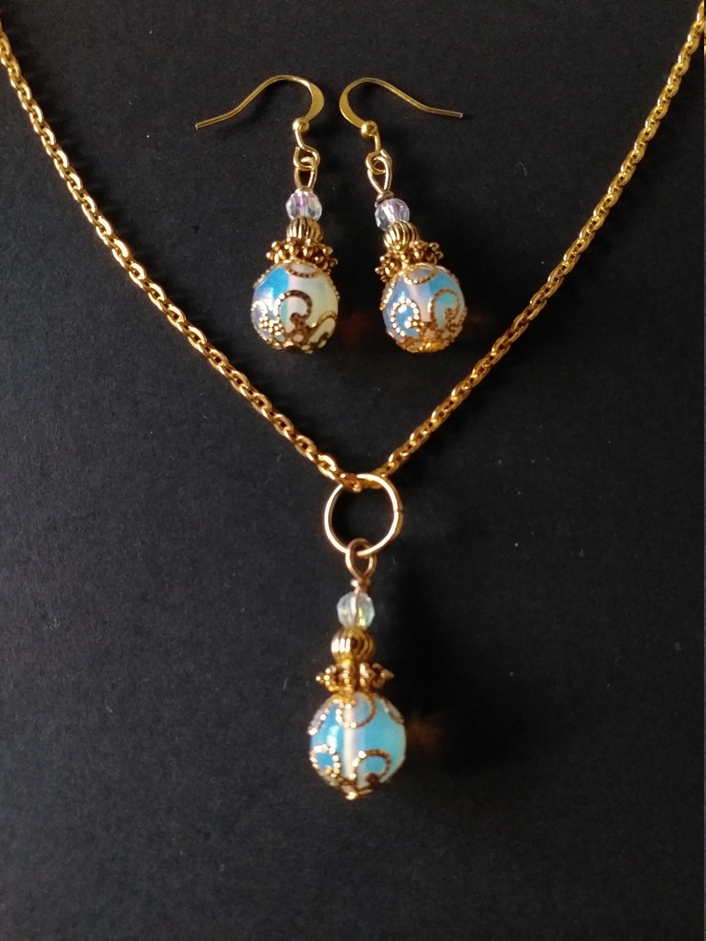 May include: A gold chain necklace with a pendant and matching earrings. The pendant and earrings feature a blue and white opal-like stone with a gold filigree design.