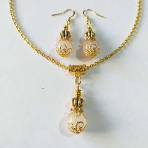 May include: A gold chain necklace with a pendant and matching earrings. The pendant and earrings feature a white, round bead with a gold filigree design.