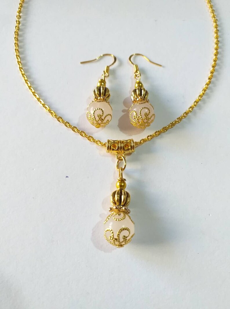 May include: A gold chain necklace with a pendant and matching earrings. The pendant and earrings feature a white, round bead with a gold filigree design.
