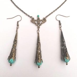 Puede incluir: Un conjunto de collar y pendientes de turquesa y bronce. El collar presenta una cadena delicada con un colgante que tiene un dise&ntilde;o de filigrana y una piedra turquesa. Los pendientes son pendientes de gota con un dise&ntilde;o de filigrana similar y piedras turquesa.