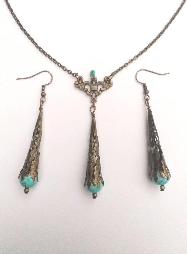 Puede incluir: Un conjunto de collar y pendientes de turquesa y bronce. El collar presenta una cadena delicada con un colgante que tiene un dise&ntilde;o de filigrana y una piedra turquesa. Los pendientes son pendientes de gota con un dise&ntilde;o de filigrana similar y piedras turquesa.