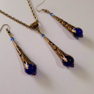 May include: A set of blue glass and gold tone earrings and necklace. The earrings and necklace feature a teardrop shaped pendant with a blue glass bead at the bottom.