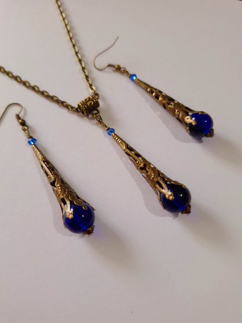 May include: A set of blue glass and gold tone earrings and necklace. The earrings and necklace feature a teardrop shaped pendant with a blue glass bead at the bottom.