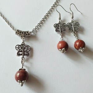 May include: A silver necklace and earrings set with a red stone pendant and matching earrings. The pendant and earrings feature a delicate butterfly design.
