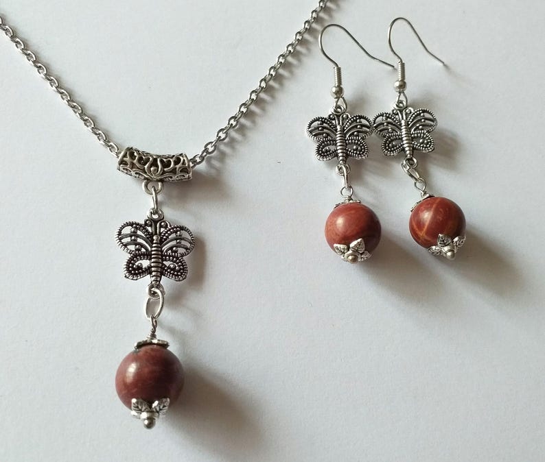 May include: A silver necklace and earrings set with a red stone pendant and matching earrings. The pendant and earrings feature a delicate butterfly design.