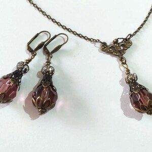 Op de afbeelding: Een set oorbellen en een ketting met een paarse glazen kraal en sierlijke bronzen metalen accenten.