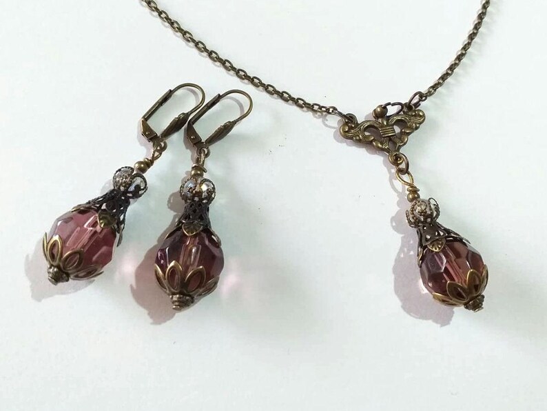 Op de afbeelding: Een set oorbellen en een ketting met een paarse glazen kraal en sierlijke bronzen metalen accenten.