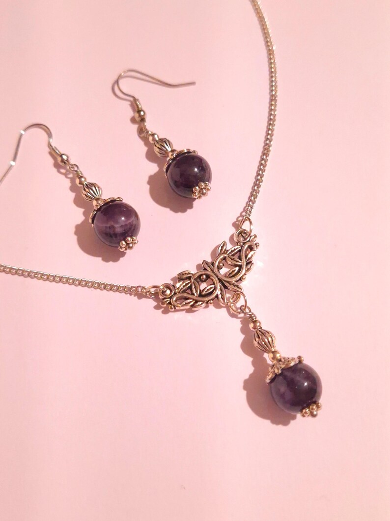 May include: A silver necklace with a delicate, ornate pendant featuring a leafy design. The pendant is accented with a round, dark purple gemstone. Matching earrings with similar gemstones dangle from silver hooks.