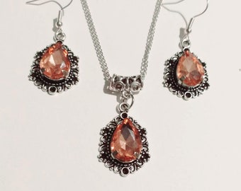 May include: A silver necklace and earrings set with a teardrop-shaped peach-colored gemstone pendant and matching earrings. The pendant and earrings are surrounded by a delicate silver filigree design.