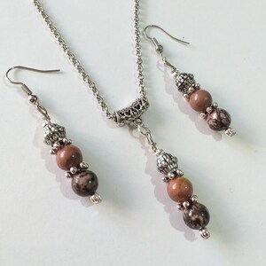 May include: A silver chain necklace with a silver pendant and three brown and black beads. Matching earrings with three brown and black beads hang from silver hooks.