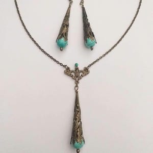 Puede incluir: Un conjunto de collar y pendientes de turquesa y bronce. El collar presenta un largo colgante adornado con una piedra turquesa en la parte inferior. Los pendientes tambi&eacute;n est&aacute;n adornados y presentan piedras turquesa.