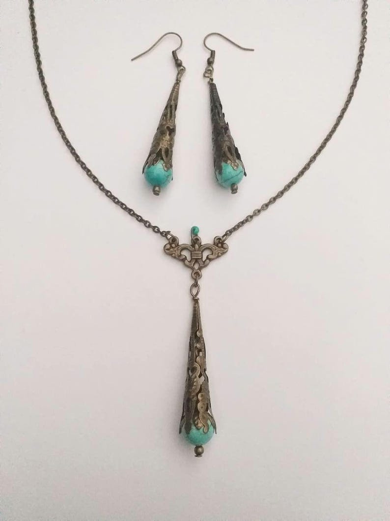 Puede incluir: Un conjunto de collar y pendientes de turquesa y bronce. El collar presenta un largo colgante adornado con una piedra turquesa en la parte inferior. Los pendientes tambi&eacute;n est&aacute;n adornados y presentan piedras turquesa.