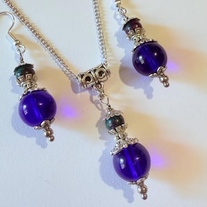 Op de afbeelding: Een sieradenset met een ketting en bijpassende oorbellen. De set bevat een zilveren ketting met een hanger en twee oorbellen. Elk stuk heeft een grote, ronde, diepblauwe kraal, geaccentueerd met zilverkleurig metaal en kleinere kralen.