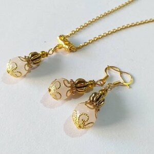 May include: A delicate gold necklace and earrings set with a pink glass bead accented with gold filigree.