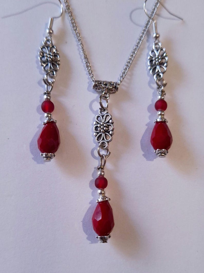Puede incluir: Un collar y pendientes de plata con cuentas rojas en forma de l&aacute;grima y detalles florales.