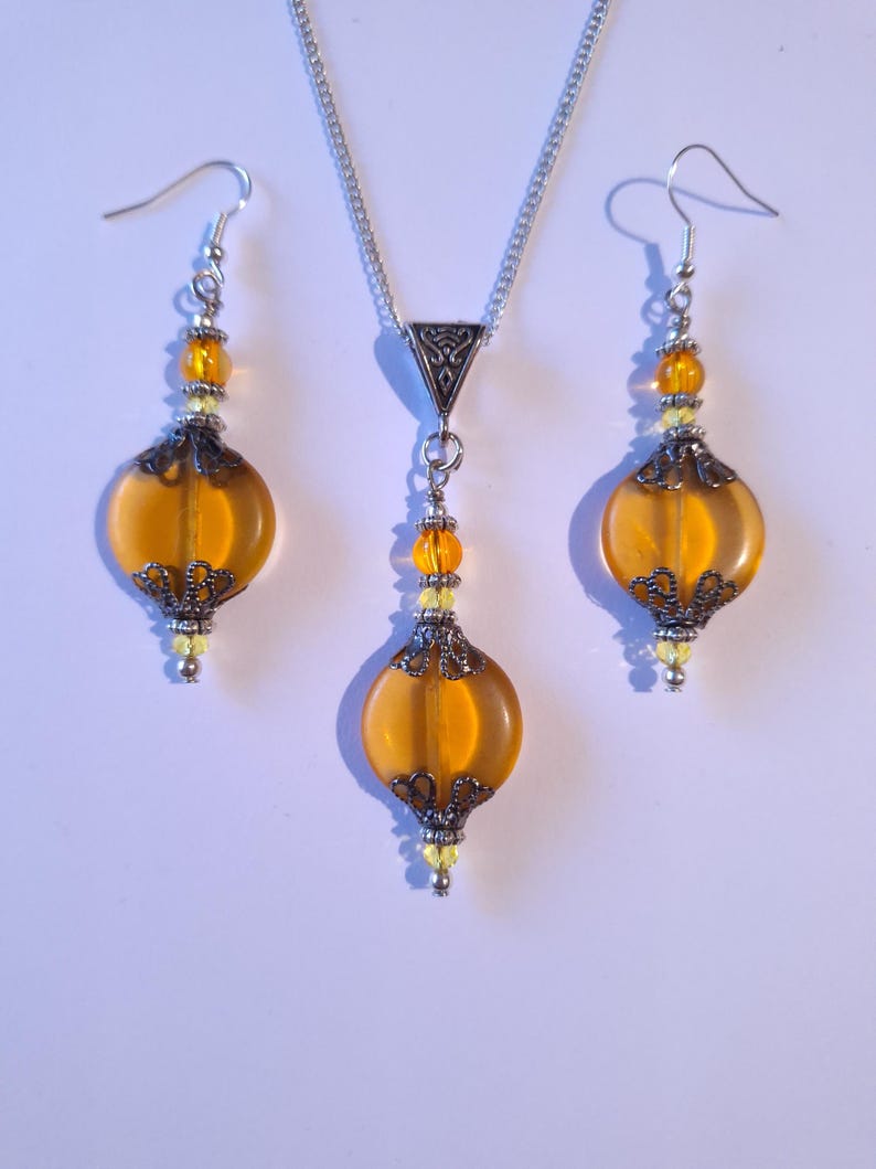 May include: A set of amber glass earrings and a necklace with silver accents. The earrings and necklace feature a round, teardrop-shaped bead with a filigree design.