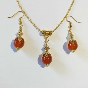 K&ouml;nnte beinhalten: Ein goldenes Halsketten- und Ohrring-Set mit einem roten Karneol-Stein-Anh&auml;nger und passenden Ohrringen. Die Halskette hat eine zarte Kette und einen kleinen goldenen Verschluss. Die Ohrringe sind Ohrh&auml;nger mit einer kleinen goldenen Perle oben.