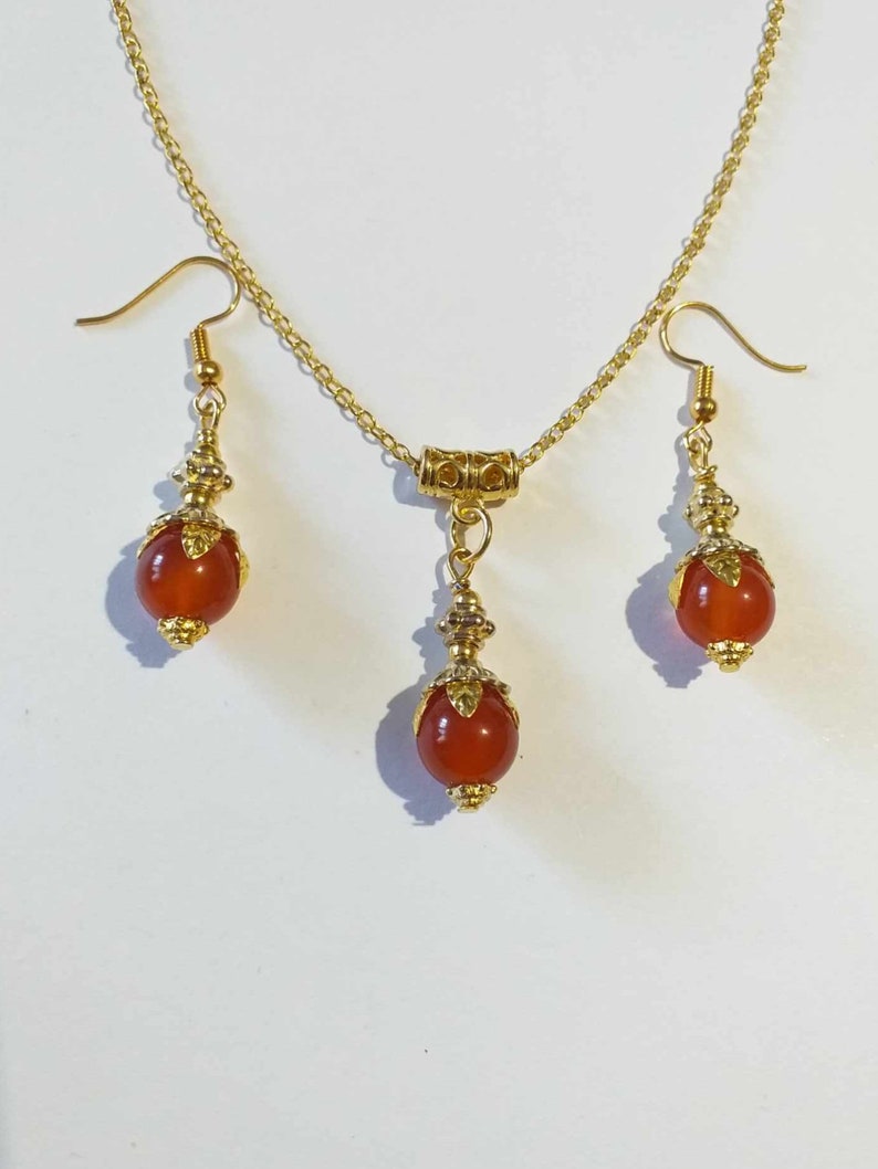 K&ouml;nnte beinhalten: Ein goldenes Halsketten- und Ohrring-Set mit einem roten Karneol-Stein-Anh&auml;nger und passenden Ohrringen. Die Halskette hat eine zarte Kette und einen kleinen goldenen Verschluss. Die Ohrringe sind Ohrh&auml;nger mit einer kleinen goldenen Perle oben.