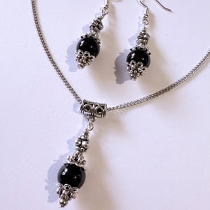 May include: A silver-toned necklace and earring set. The necklace features a pendant with a black bead and decorative silver accents. The earrings have a similar design, with black beads and silver-colored metalwork. The jewelry set is displayed on a white surface.