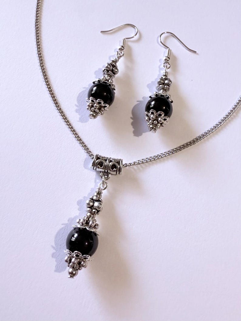 May include: A silver-toned necklace and earring set. The necklace features a pendant with a black bead and decorative silver accents. The earrings have a similar design, with black beads and silver-colored metalwork. The jewelry set is displayed on a white surface.