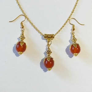 K&ouml;nnte beinhalten: Ein goldenes Halsketten- und Ohrring-Set mit roten Karneol-Steinen. Die Halskette hat eine goldene Kette und einen kleinen goldenen Anh&auml;nger mit einem Karneol-Stein. Die Ohrringe sind Ohrh&auml;nger mit Karneol-Steinen.