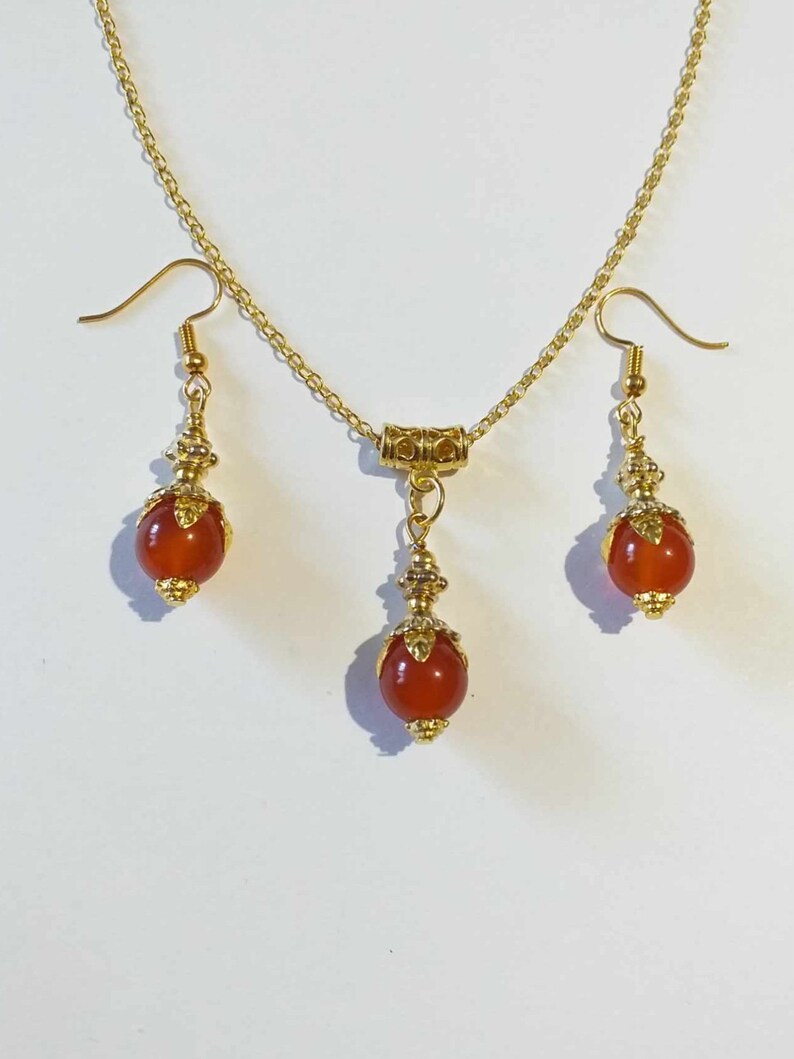K&ouml;nnte beinhalten: Ein goldenes Halsketten- und Ohrring-Set mit roten Karneol-Steinen. Die Halskette hat eine goldene Kette und einen kleinen goldenen Anh&auml;nger mit einem Karneol-Stein. Die Ohrringe sind Ohrh&auml;nger mit Karneol-Steinen.