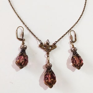 Op de afbeelding: Een set sieraden met een delicate gouden ketting met een hanger en bijpassende oorbellen. De hanger en oorbellen hebben elk een traanvormig paars kristal met ingewikkelde gouden accenten.