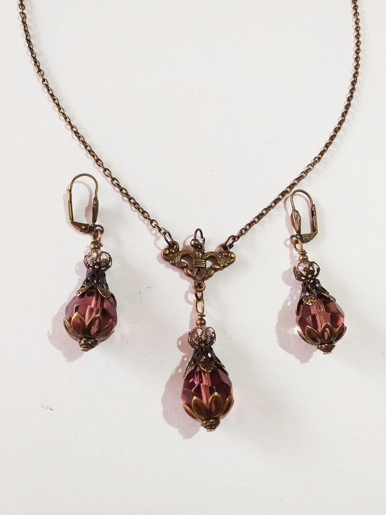 Op de afbeelding: Een set sieraden met een delicate gouden ketting met een hanger en bijpassende oorbellen. De hanger en oorbellen hebben elk een traanvormig paars kristal met ingewikkelde gouden accenten.