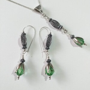 May include: A silver necklace with a pendant featuring a green crystal and ornate silver accents. Matching earrings with green crystals and silver accents.