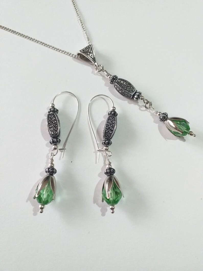 May include: A silver necklace with a pendant featuring a green crystal and ornate silver accents. Matching earrings with green crystals and silver accents.