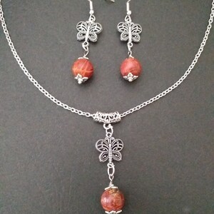 May include: A silver chain necklace with a red and brown stone pendant and a silver butterfly charm. Matching earrings with red and brown stone drops and silver butterfly charms.