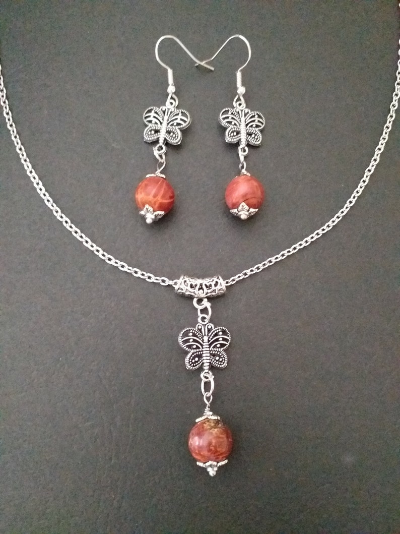 May include: A silver chain necklace with a red and brown stone pendant and a silver butterfly charm. Matching earrings with red and brown stone drops and silver butterfly charms.