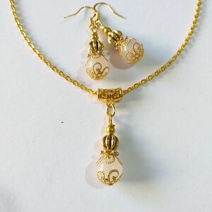May include: A gold chain necklace with a white pendant and matching earrings. The pendant and earrings feature a gold filigree design and a white stone.