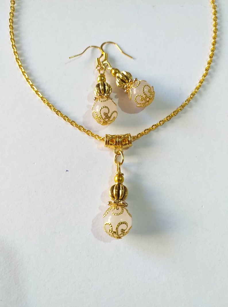 May include: A gold chain necklace with a white pendant and matching earrings. The pendant and earrings feature a gold filigree design and a white stone.