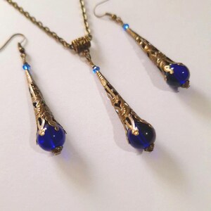 May include: A gold necklace and earrings set with blue glass teardrop pendants. The necklace has a chain and a small decorative piece at the top. The earrings are dangle earrings with a hook closure.