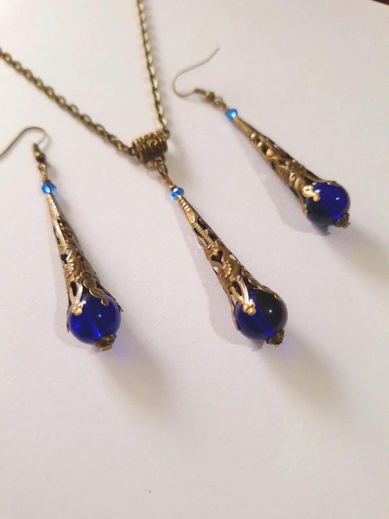 May include: A gold necklace and earrings set with blue glass teardrop pendants. The necklace has a chain and a small decorative piece at the top. The earrings are dangle earrings with a hook closure.