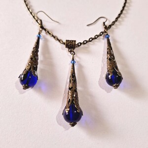 May include: A set of blue glass and bronze jewelry featuring a necklace and earrings. The necklace has a chain with a pendant that is a teardrop shape with a blue glass bead at the bottom. The earrings are also teardrop shaped with blue glass beads at the bottom.