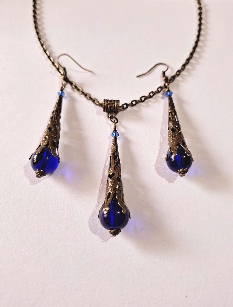 May include: A set of blue glass and bronze jewelry featuring a necklace and earrings. The necklace has a chain with a pendant that is a teardrop shape with a blue glass bead at the bottom. The earrings are also teardrop shaped with blue glass beads at the bottom.