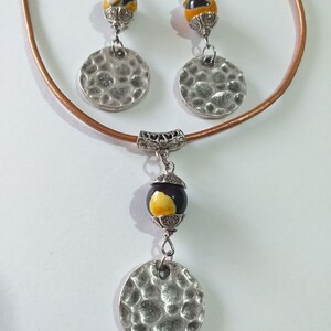 May include: A jewelry set featuring a necklace and earrings. The necklace has a brown leather cord and a pendant with a black and yellow bead and a hammered silver disc. The earrings have matching beads and discs. The set is designed with silver metal accents.