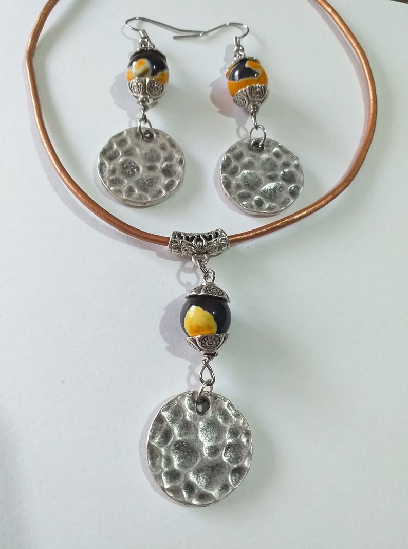 May include: A jewelry set featuring a necklace and earrings. The necklace has a brown leather cord and a pendant with a black and yellow bead and a hammered silver disc. The earrings have matching beads and discs. The set is designed with silver metal accents.