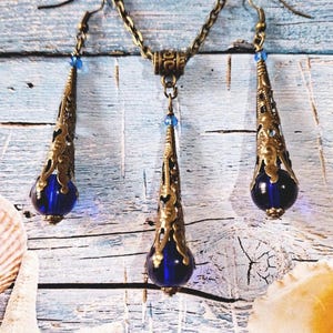 May include: A gold necklace and earrings set with blue glass teardrop pendants. The necklace has a chain link design and the earrings have a hook closure.