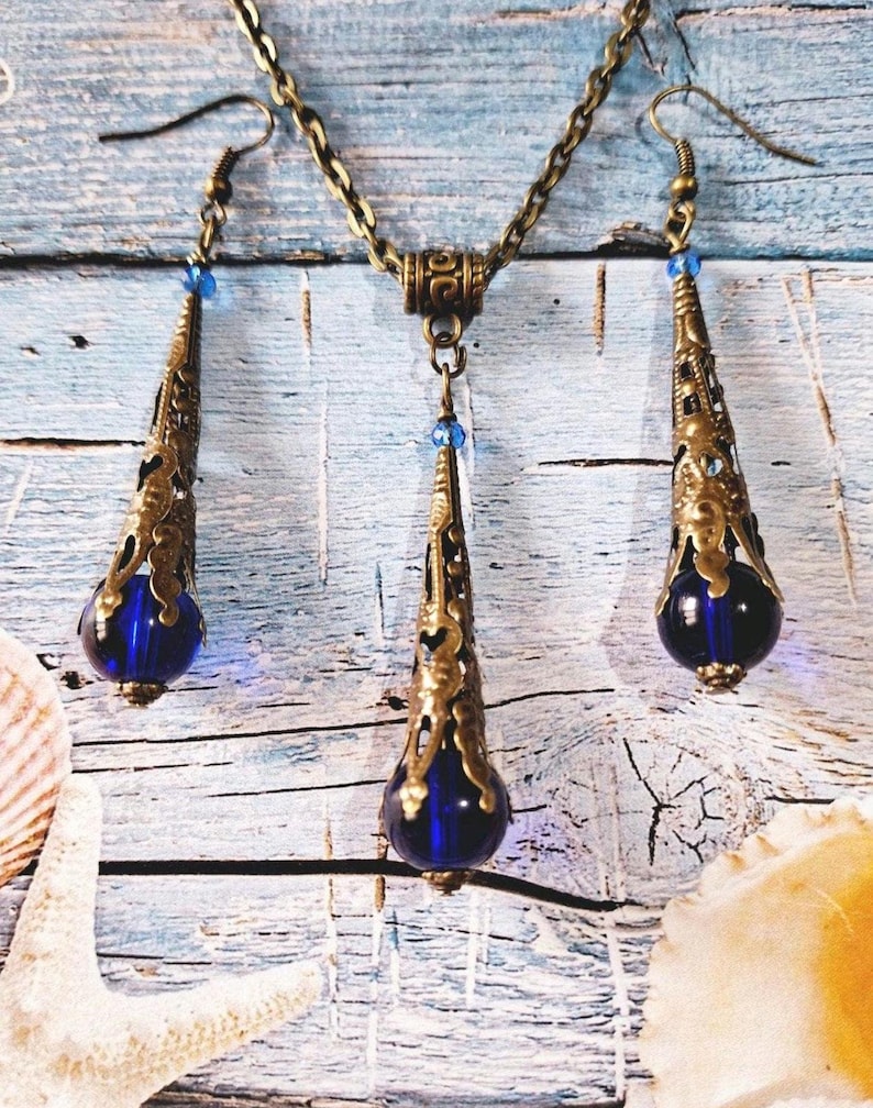 May include: A gold necklace and earrings set with blue glass teardrop pendants. The necklace has a chain link design and the earrings have a hook closure.