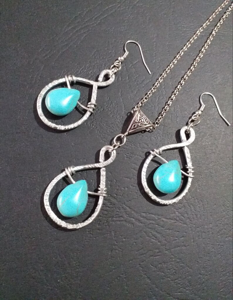 K&ouml;nnte beinhalten: Ein silbernes Halsketten- und Ohrring-Set mit t&uuml;rkisfarbenen Tr&auml;nensteinen. Die Halskette hat einen dreieckigen Anh&auml;nger mit einem keltischen Knotendesign. Die Ohrringe sind im H&auml;nge-Stil mit einem tropfenf&ouml;rmigen Stein.