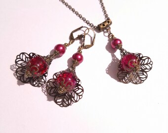 Könnte beinhalten: Ein Set aus Ohrringen und einer Halskette mit floralem Design. Die Ohrringe und die Halskette haben eine rote Glasperle mit glitzernder Oberfläche und einem filigranen Design aus Bronze. Die Halskette hat eine Kette aus Bronze.