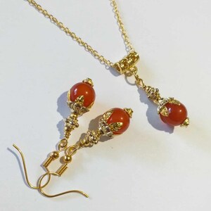 K&ouml;nnte beinhalten: Ein goldenes Halsketten- und Ohrring-Set mit orangefarbenen Edelsteinperlen und goldenen Akzenten. Die Halskette hat eine Gliederkette und einen Karabinerverschluss. Die Ohrringe sind Tropfenform mit einer kleinen &Ouml;se f&uuml;r Ohrl&ouml;cher.