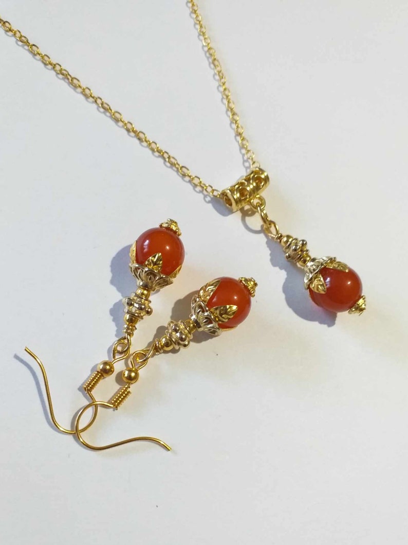 K&ouml;nnte beinhalten: Ein goldenes Halsketten- und Ohrring-Set mit orangefarbenen Edelsteinperlen und goldenen Akzenten. Die Halskette hat eine Gliederkette und einen Karabinerverschluss. Die Ohrringe sind Tropfenform mit einer kleinen &Ouml;se f&uuml;r Ohrl&ouml;cher.
