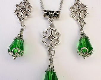 Puede incluir: Un conjunto de joyería compuesto por un collar y dos pendientes a juego. Cada pieza presenta un cristal facetado en forma de lágrima de color verde esmeralda, engastado en filigrana de metal plateado y suspendido de un conector plateado.