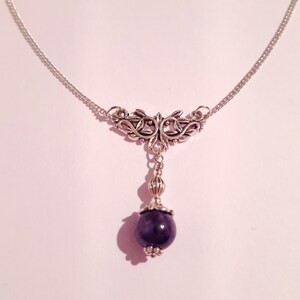 Puede incluir: Un collar de cadena de plata con un delicado colgante de dise&ntilde;o floral y una cuenta de piedra preciosa de color p&uacute;rpura oscuro.