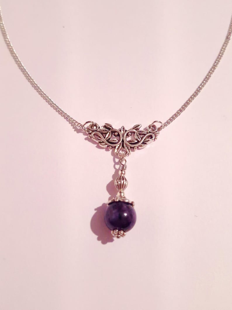 Puede incluir: Un collar de cadena de plata con un delicado colgante de dise&ntilde;o floral y una cuenta de piedra preciosa de color p&uacute;rpura oscuro.
